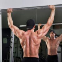 A topless man doing a shoulder press while looking in the mirror
