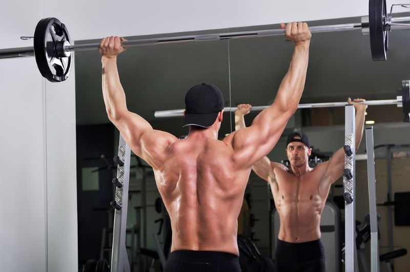 A topless man doing a shoulder press while looking in the mirror