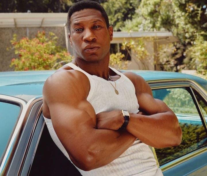 Jonathan majors leaning on a car showing off his bulky biceps